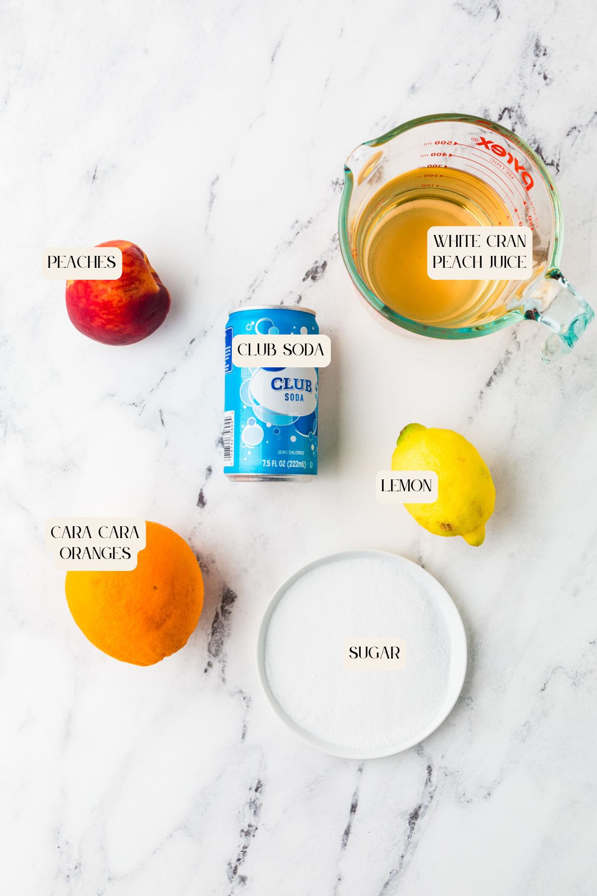 Ingredients for a white sangria mocktail are arranged on a marble surface: a peach, Cara Cara orange, lemon, club soda, measuring cup of white cran-peach juice, and a plate of sugar, each labeled.