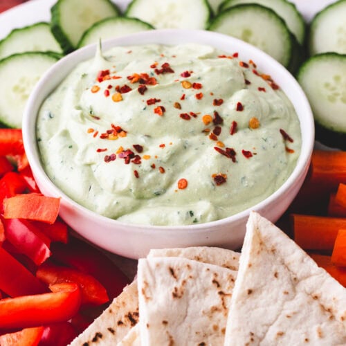 A white bowl of creamy dip topped with red pepper flakes is surrounded by sliced cucumbers, red bell peppers, carrot sticks, and pieces of flatbread on a white plate.