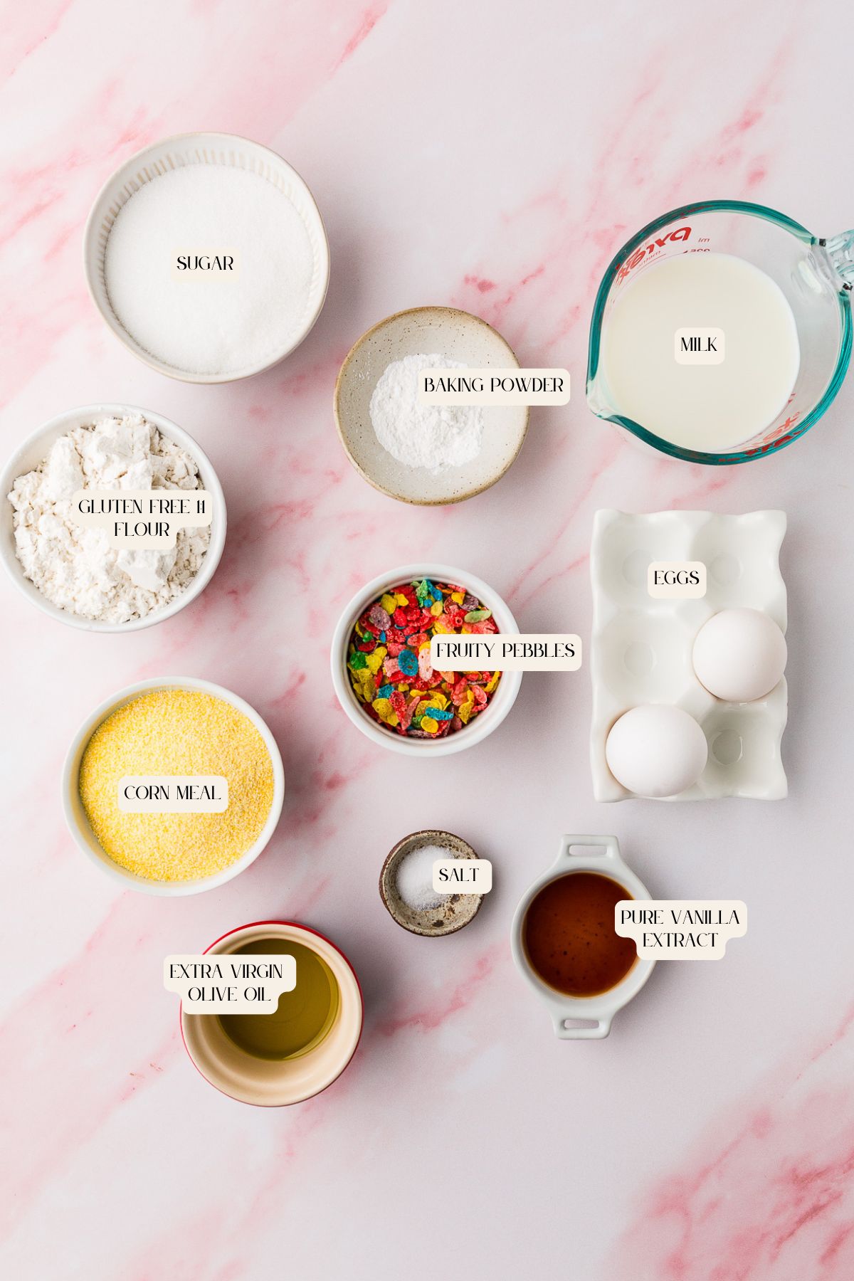 A flat lay of labeled ingredients for Fruity Pebble Waffles on a pink surface, including sugar, gluten-free flour, corn meal, extra virgin olive oil, baking powder, Fruity Pebbles cereal, salt, vanilla extract, milk, and two eggs.