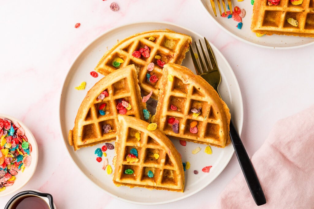 A round Fruity Pebble Waffle cut into quarters is served on a white plate with a black fork. Its topped with colorful cereal pieces, with more scattered nearby. A syrup pitcher and another plate are partially visible.
