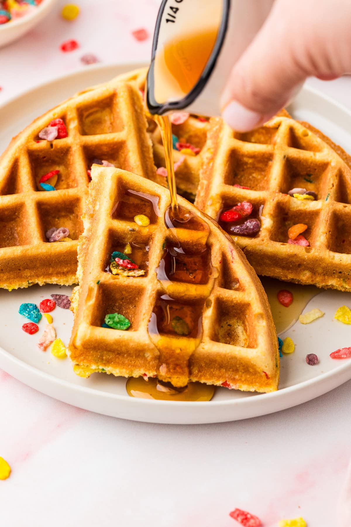 A hand pours syrup over Fruity Pebble Waffles on a white plate, topped with vibrant cereal pieces. Some cereal bits are scattered around the plate on a pink surface.