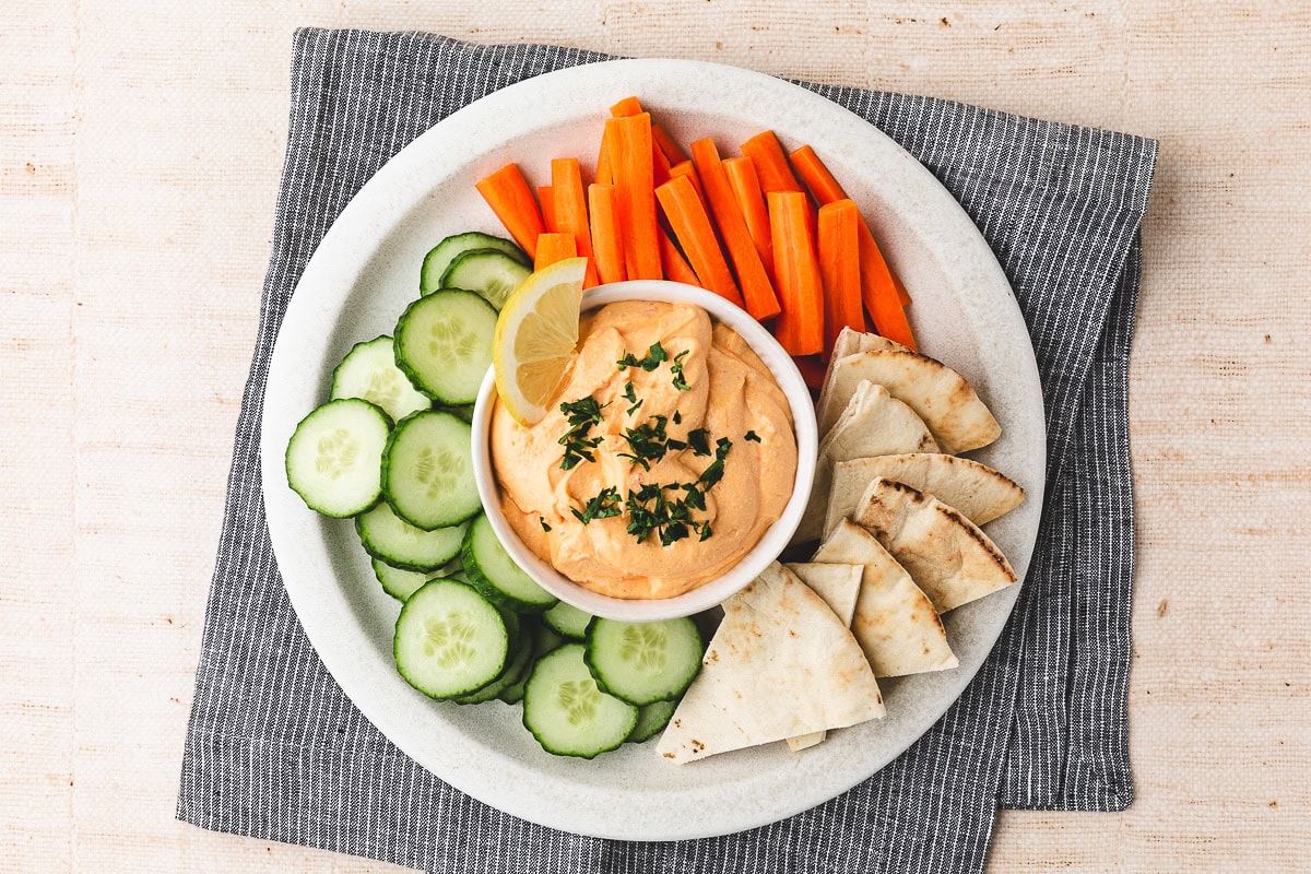 A plate with sliced cucumbers, carrot sticks, pita wedges, and a bowl of harissa cottage cheese dip garnished with herbs and a lemon slice, set on a gray napkin.