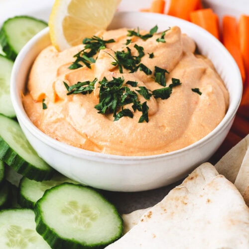 A bowl of creamy harissa cottage cheese dip topped with chopped herbs and a lemon wedge, surrounded by sliced cucumbers, carrot sticks, and pieces of pita bread on a white plate.