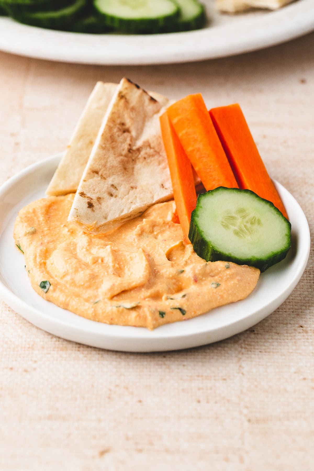 A small plate with a swirl of hummus, pita bread triangles, carrot sticks, and a cucumber slice sits alongside harissa cottage cheese dip on a light-colored surface.