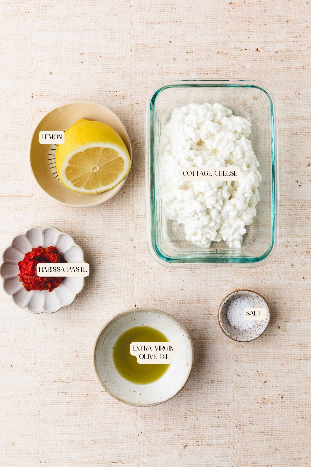 Five ingredients on a light textured surface: half a lemon, cottage cheese in a glass container, a small bowl of harissa paste, olive oil, and salt—each labeled and ready to make a vibrant harissa cottage cheese dip.