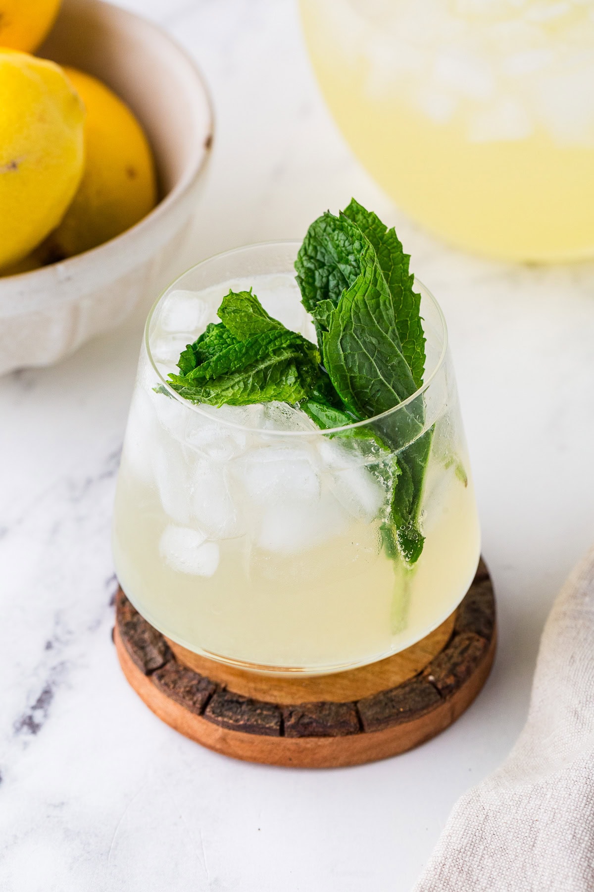 A glass of sparkling lemonade mocktail garnished with fresh mint leaves sits on a wooden coaster beside a bowl of lemons on a white marble surface.