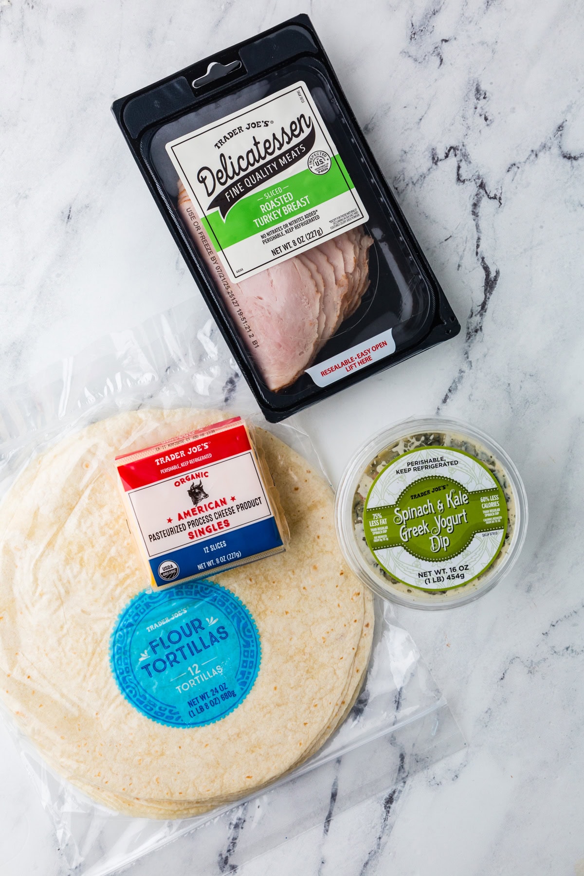 Four Trader Joe’s products on a marble surface: a package of flour tortillas, a pack of sliced oven roasted turkey breast, a container of spinach & kale Greek yogurt dip, and a block of American cheese.