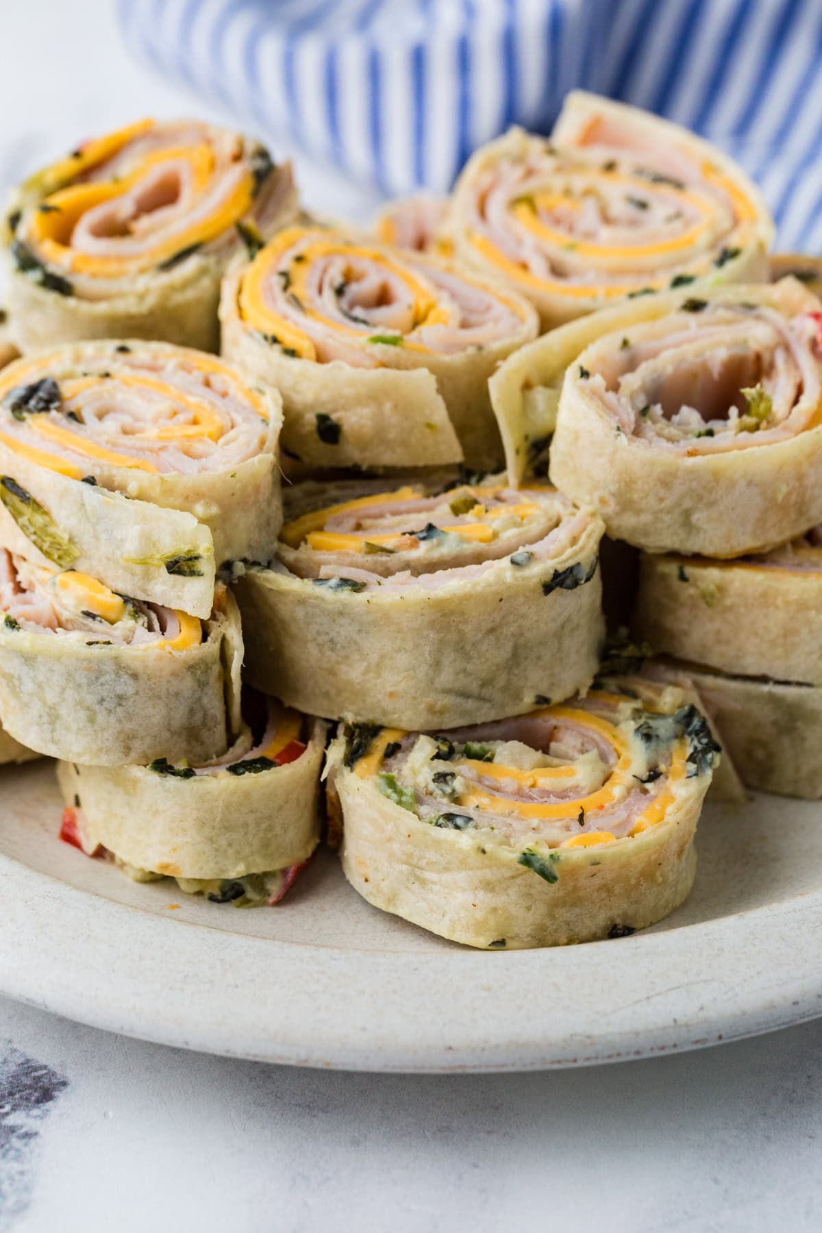 A plate of rolled tortilla pinwheel sandwiches filled with slices of turkey, cheddar cheese, and greens, arranged in a neat stack. A blue and white striped cloth is in the background.