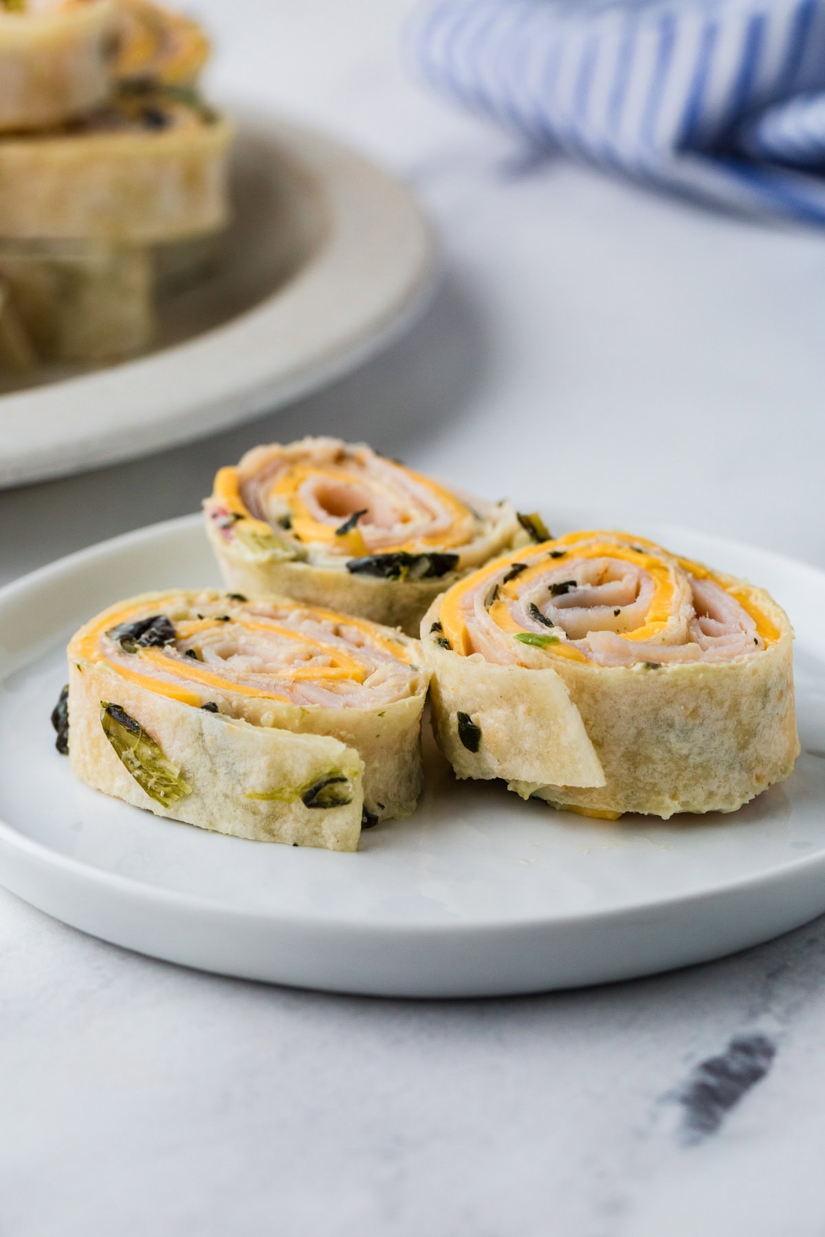 Three tortilla pinwheels with visible turkey, cheese, and spinach filling are arranged on a white plate, with more pinwheels blurred in the background.