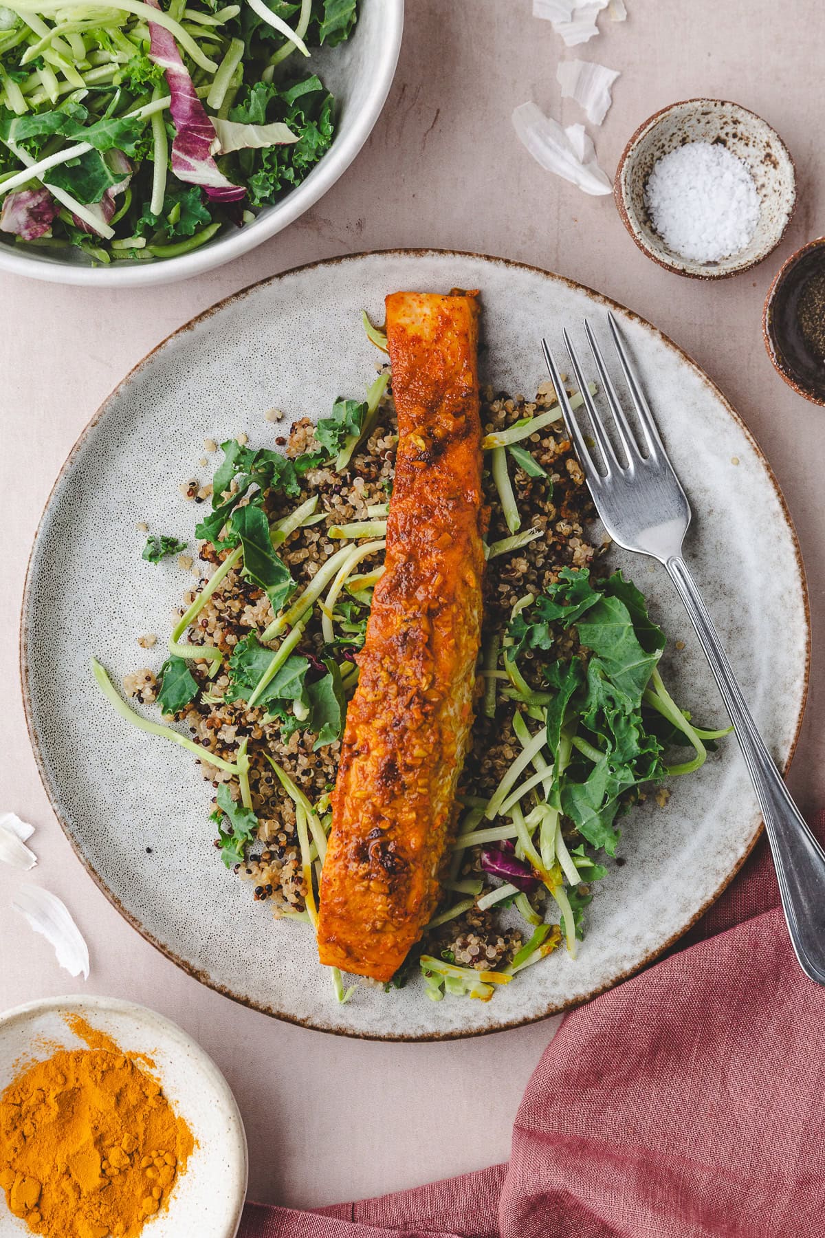 A plate with a baked salmon fillet on top of quinoa, greens, and julienned vegetables, with a fork on the side. Nearby are small bowls of salt and turmeric, and a bowl of salad greens.