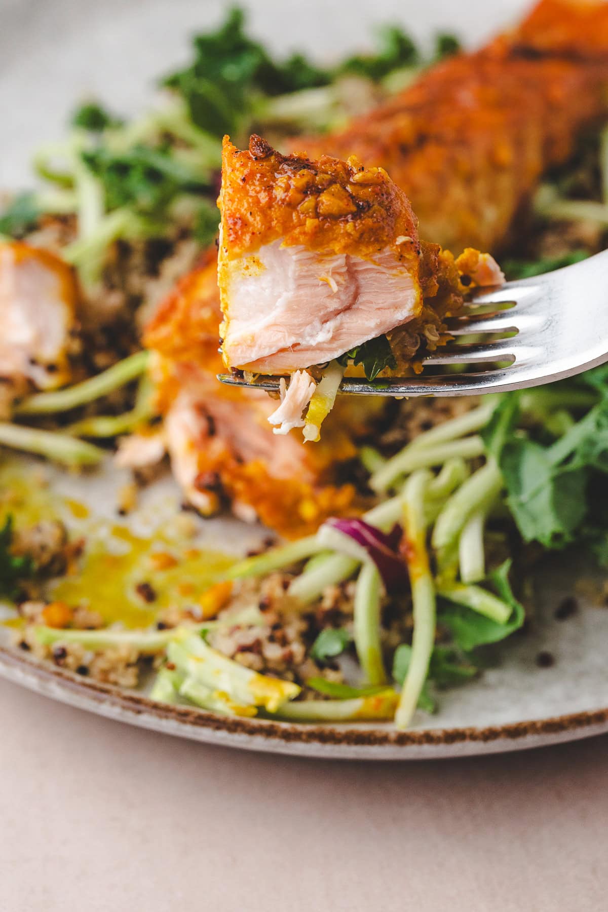 A close-up of a fork holding a bite of baked salmon with a golden spice crust, above a plate of quinoa salad mixed with greens and shredded vegetables.