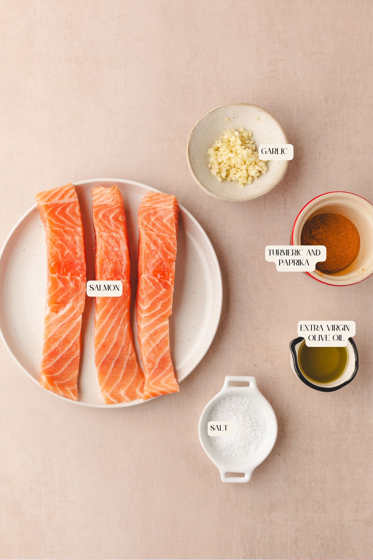 Three raw salmon fillets on a plate, surrounded by small bowls containing minced garlic, turmeric and paprika, extra virgin olive oil, and salt, all labeled on a light beige background.