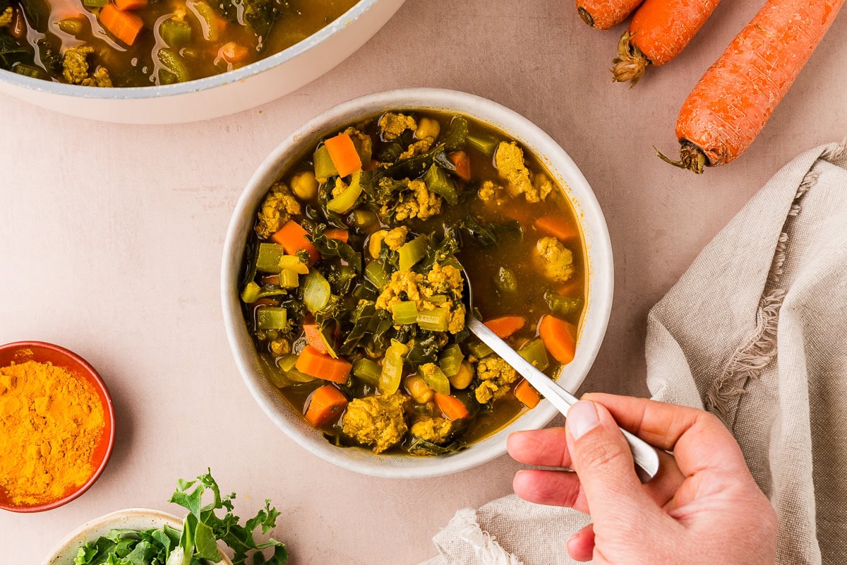 A hand holds a spoon in a bowl of vegetable soup with carrots, greens, and broth on a beige table, surrounded by fresh carrots, herbs, and a bowl of turmeric.