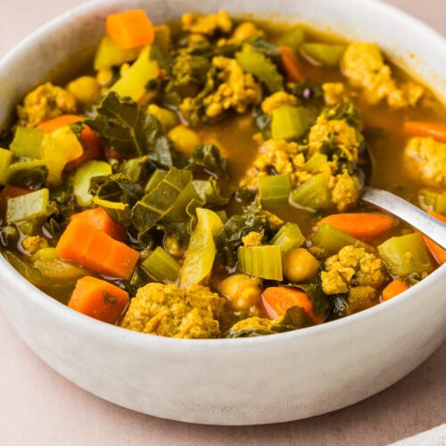 A bowl of hearty vegetable soup filled with chunks of carrots, celery, chickpeas, leafy greens, and pieces of plant-based protein, with a spoon resting inside the bowl on a light surface.