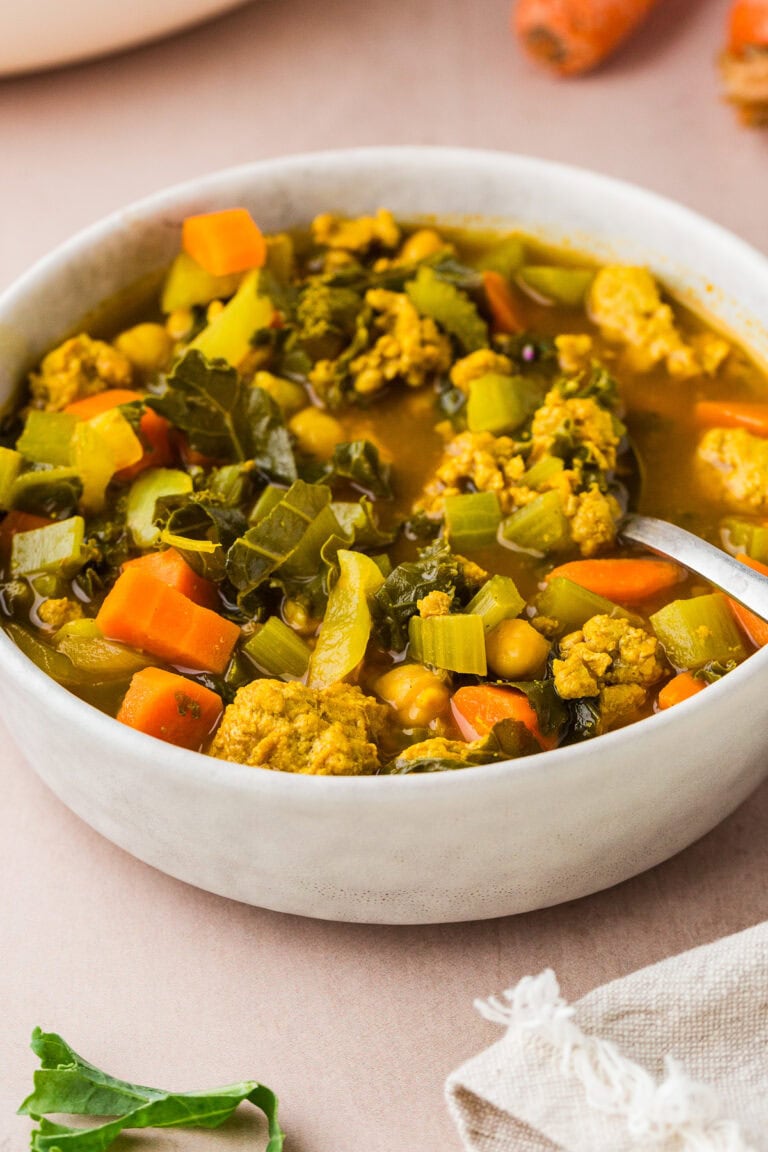 A bowl of hearty vegetable soup filled with chunks of carrots, celery, chickpeas, leafy greens, and pieces of plant-based protein, with a spoon resting inside the bowl on a light surface.