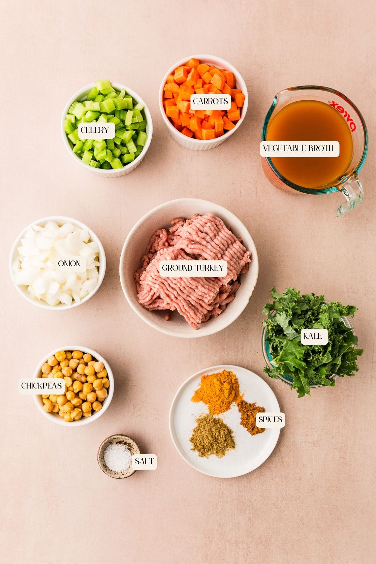 Flat lay of labeled ingredients in bowls for a soup: ground turkey, chopped celery, diced carrots, vegetable broth, chopped onion, kale, chickpeas, salt, and a plate with various spices on a light background.