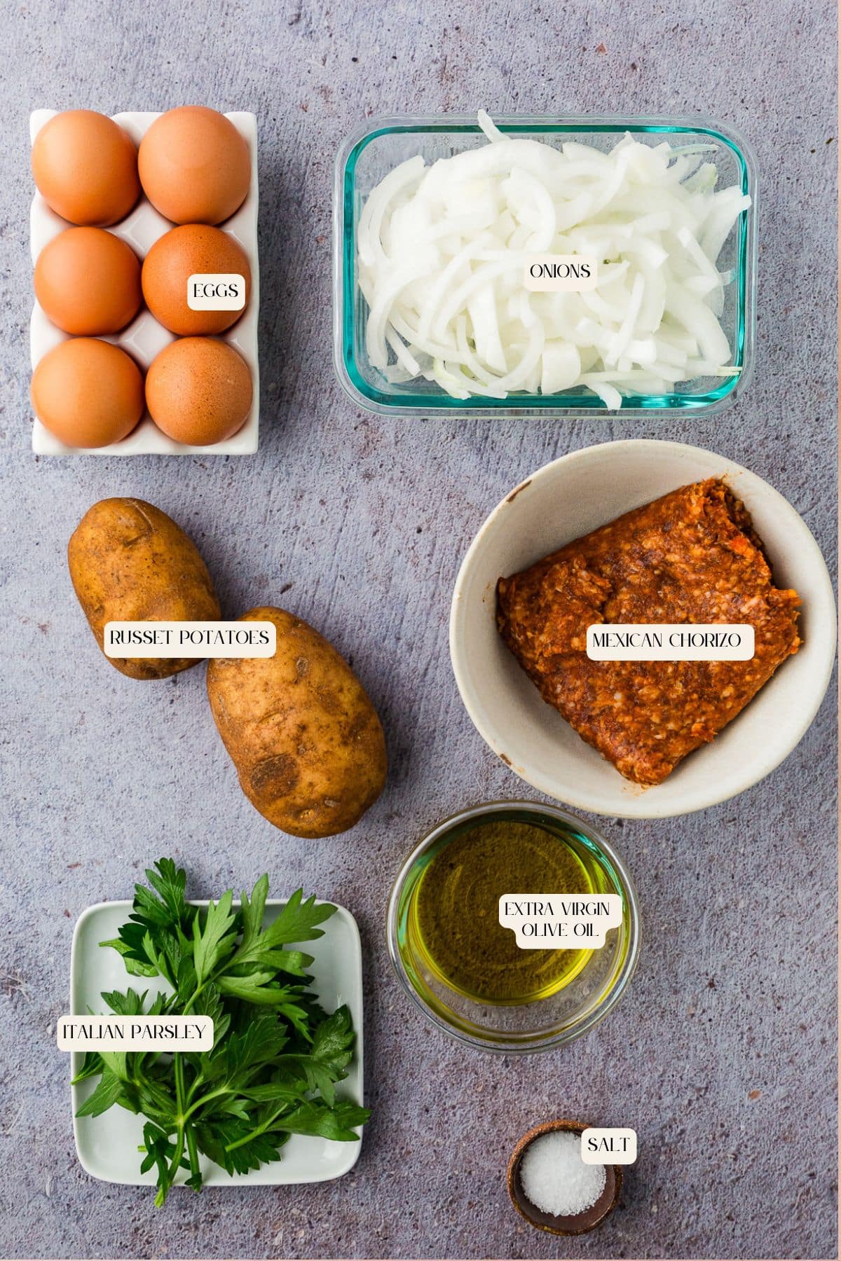 A flat lay of ingredients labeled: eggs, sliced onions, russet potatoes, Italian parsley, Mexican chorizo, extra virgin olive oil, and salt on a gray surface—perfect for cooking creative meals during things to do in a power outage.