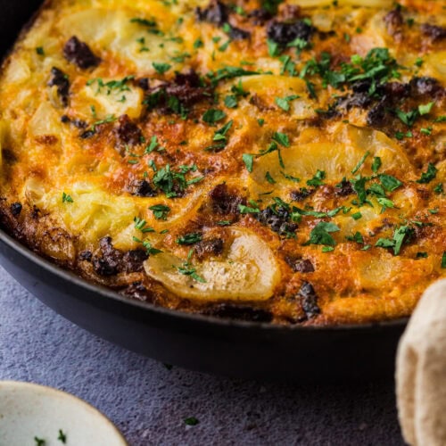 A close-up of a golden-brown spanish tortilla with chorizo in a black skillet, topped with chopped herbs—perfect comfort food and one of the best things to do in a power outage. A small bowl of fresh herbs and a beige cloth are nearby on a textured surface.
