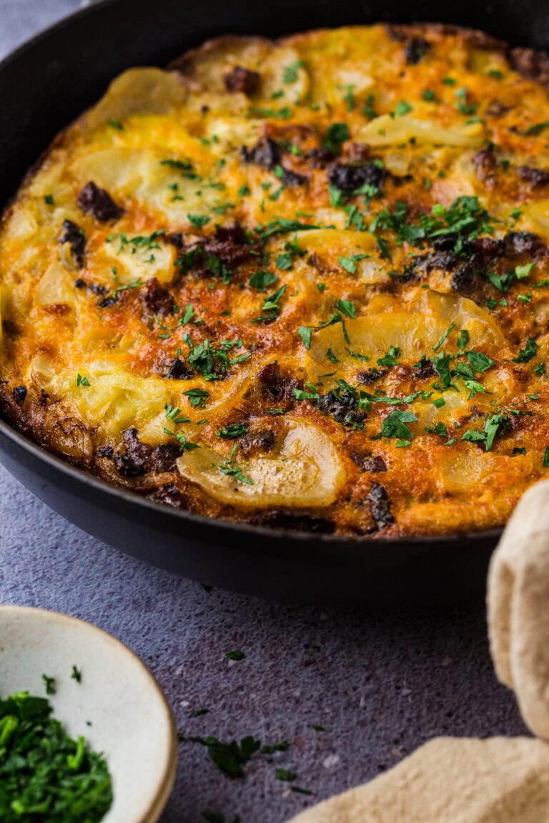 A close-up of a golden-brown spanish tortilla with chorizo in a black skillet, topped with chopped herbs—perfect comfort food and one of the best things to do in a power outage. A small bowl of fresh herbs and a beige cloth are nearby on a textured surface.