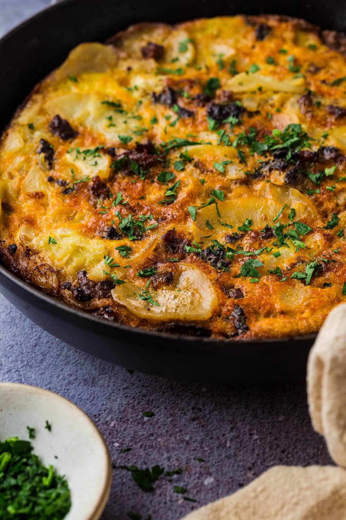 A close-up of a frittata in a skillet, topped with chopped fresh herbs—an ideal meal for things to do in a power outage. Slices of potato and browned sausage are visible, with a small bowl of herbs and a beige napkin beside the skillet.
