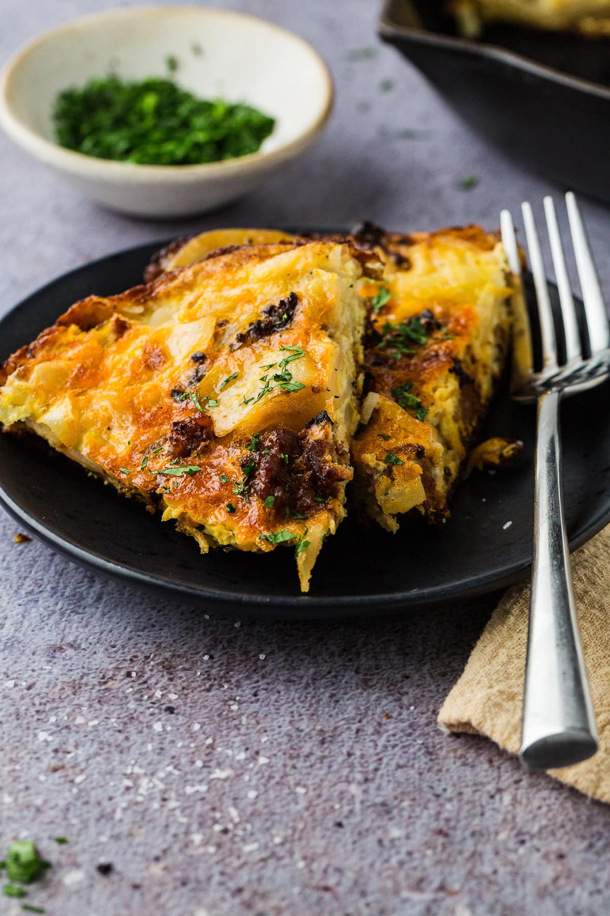 A close-up of two slices of spanish tortilla with chorizo with herbs on a black plate, next to a fork and a bowl of chopped parsley—perfect comfort food when looking for things to do in a power outage, on a light textured surface.