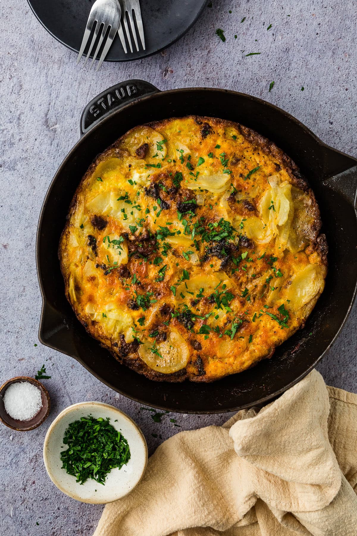 A baked frittata with golden-brown edges, sliced potatoes, herbs, and cheese sits in a black skillet—a perfect meal idea for things to do in a power outage. Nearby are chopped parsley, salt, a beige cloth, and two forks on a black plate.