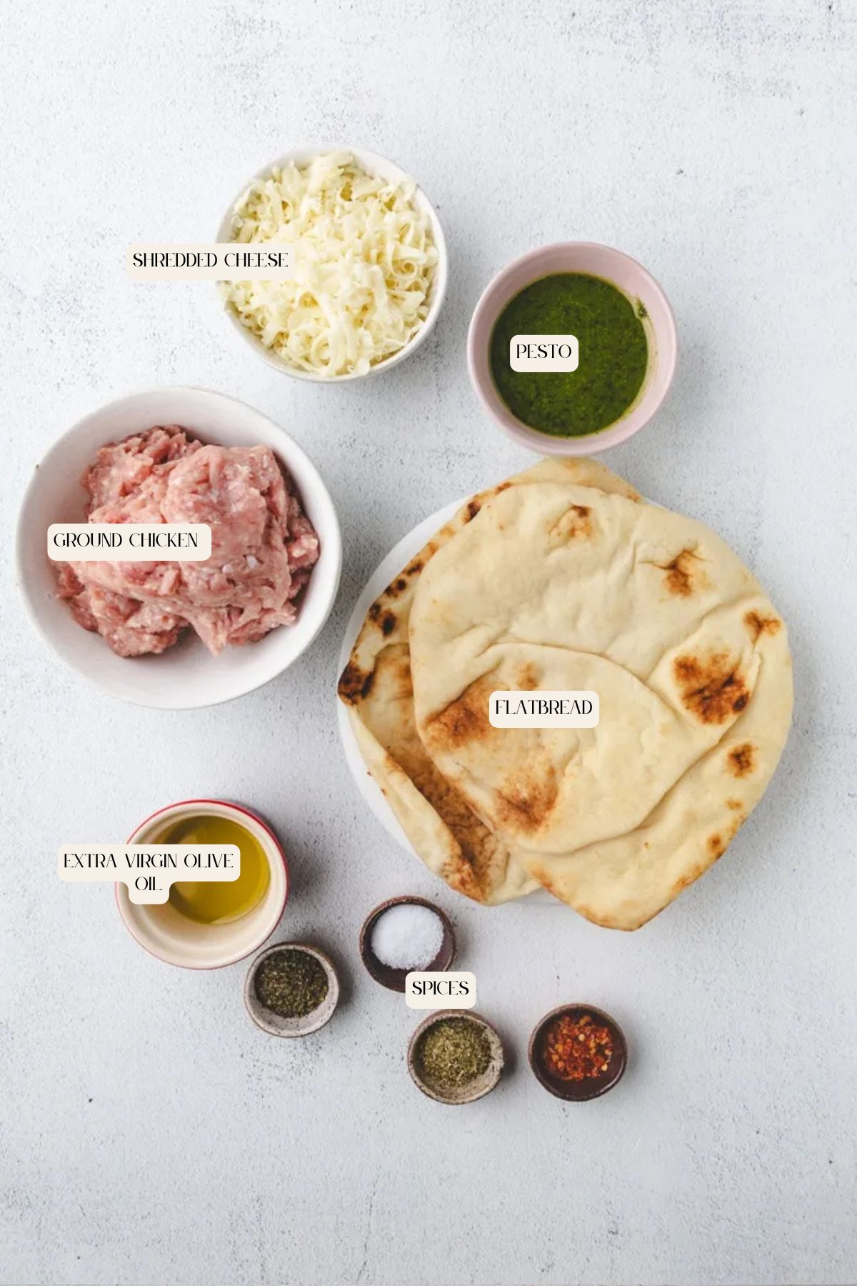 A flat lay of labeled ingredients for chicken pesto flatbread: shredded cheese, pesto, ground chicken, flatbread, extra virgin olive oil, and three small bowls of spices, all arranged on a light gray surface.