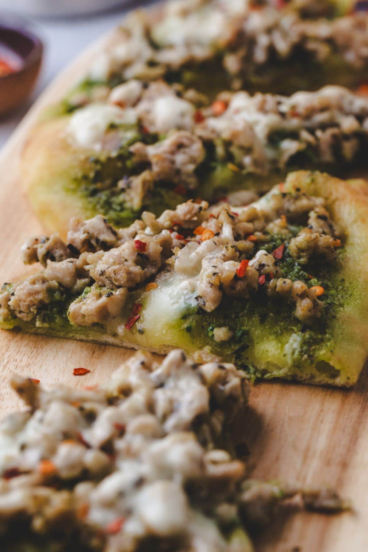 A close-up of chicken pesto flatbread slices topped with seasoned ground chicken, melted cheese, and red pepper flakes on a wooden board.