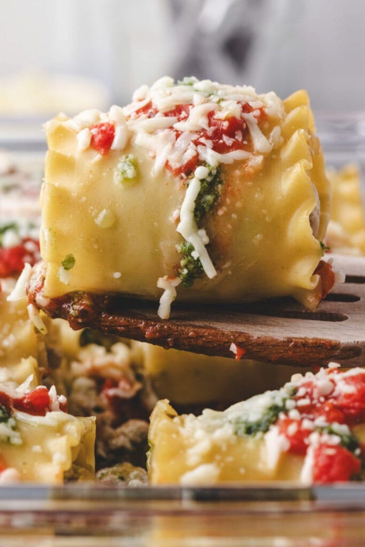 A close-up of a chicken pesto lasagna roll up filled with spinach and ricotta, topped with tomato sauce and shredded cheese, being lifted from a glass baking dish with a wooden spatula.