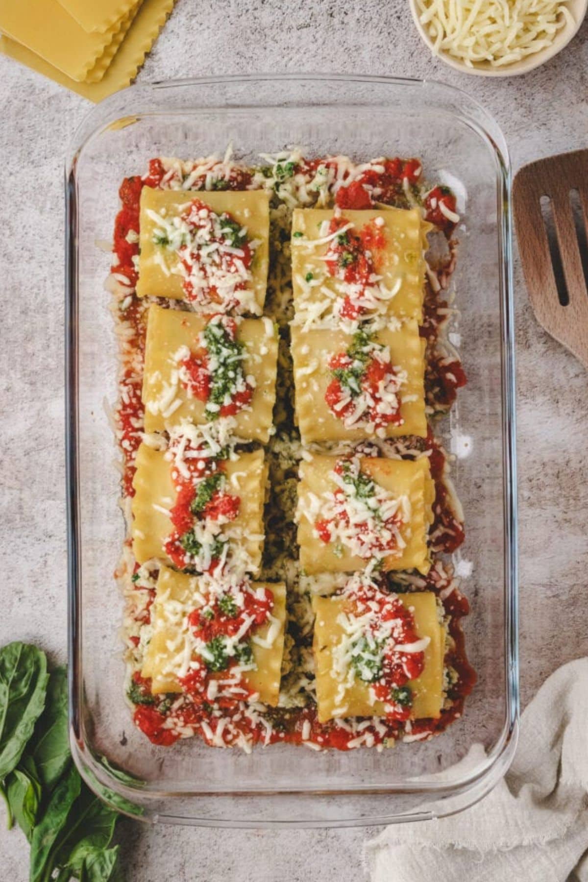 A glass baking dish filled with chicken pesto lasagna roll ups, topped with tomato sauce, shredded cheese, and green pesto, sits on a light surface next to fresh basil, a wooden spatula, and a bowl of shredded cheese.