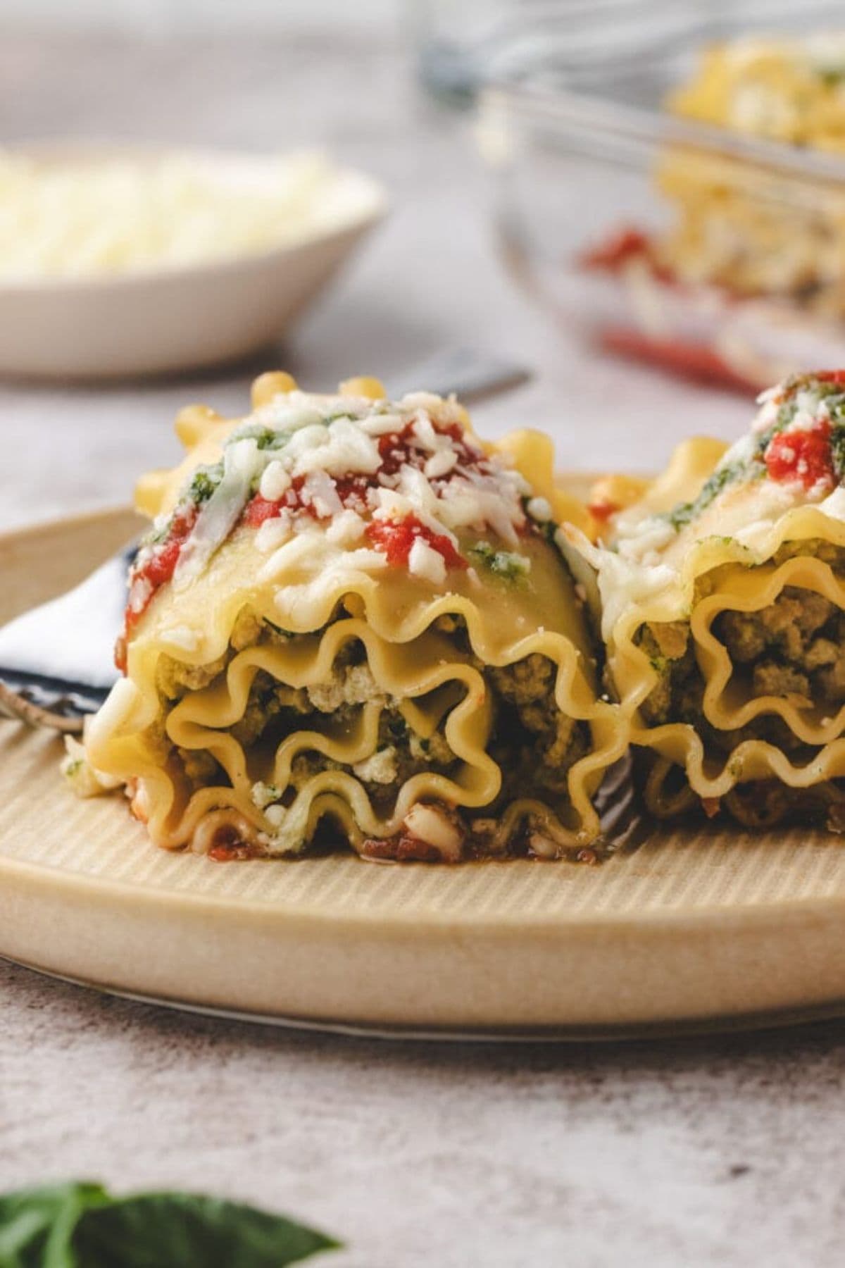 Two rolled lasagna noodles filled with spinach and cheese, inspired by chicken pesto lasagna roll ups, topped with tomato sauce and melted cheese, served on a beige plate. A fork and a bowl of shredded cheese are blurred in the background.