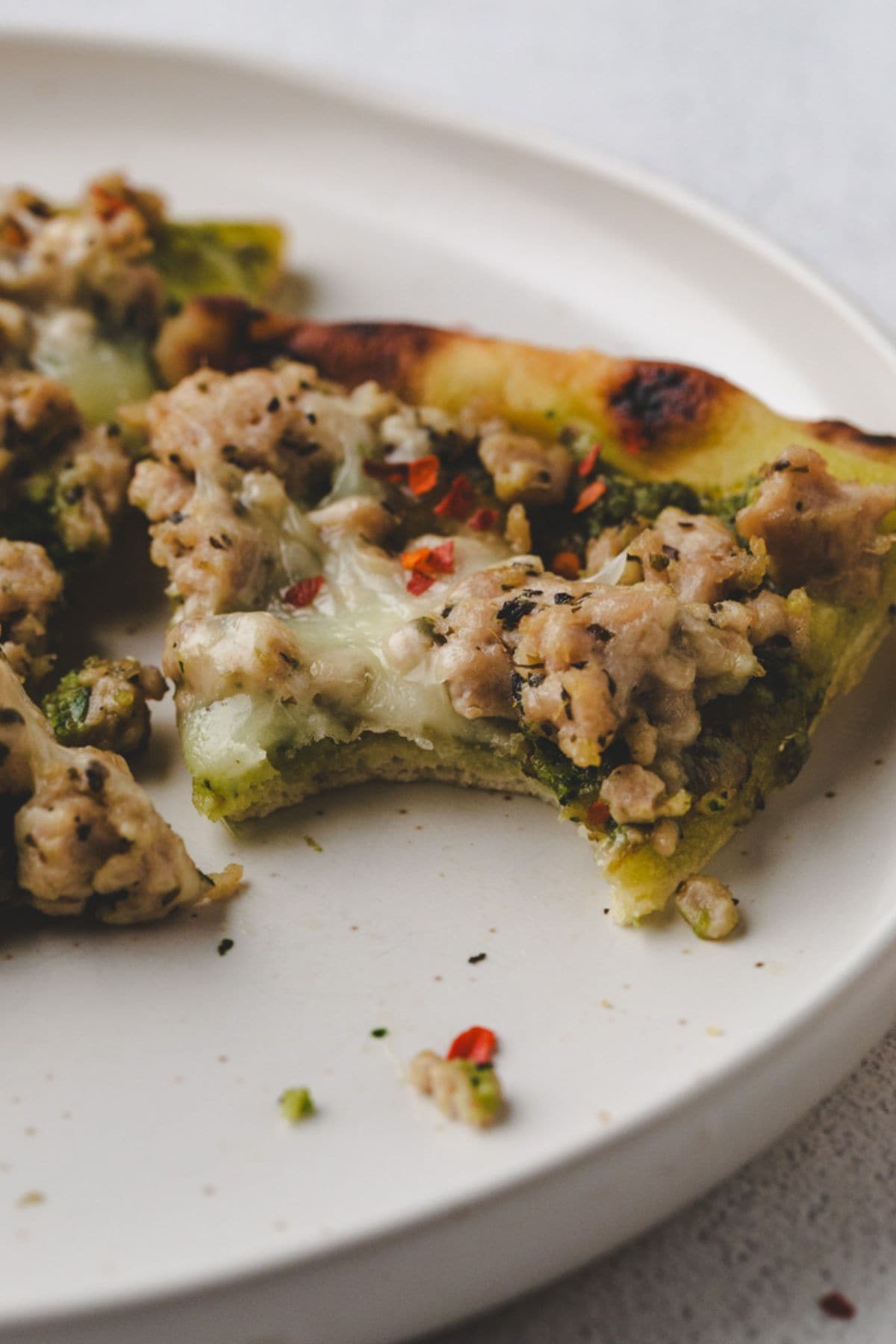 A close-up of a chicken pesto flatbread pizza slice with a bite taken out, topped with ground meat, melted cheese, and herbs, served on a white plate.