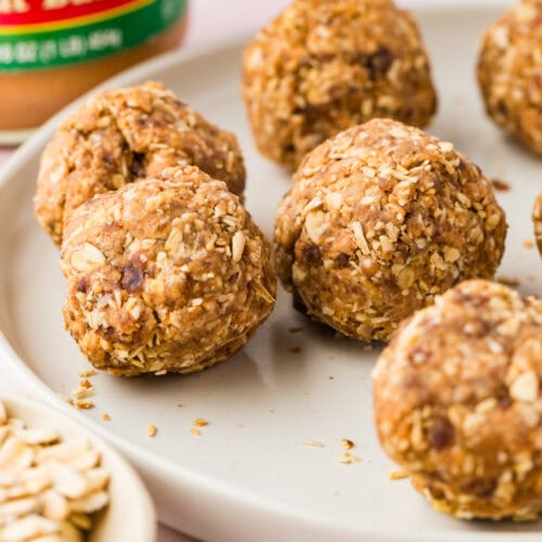 Several oat and peanut butter and date protein balls rest on a white plate. A jar of peanut butter and a small bowl of oats are visible in the background. The energy balls appear textured and homemade.