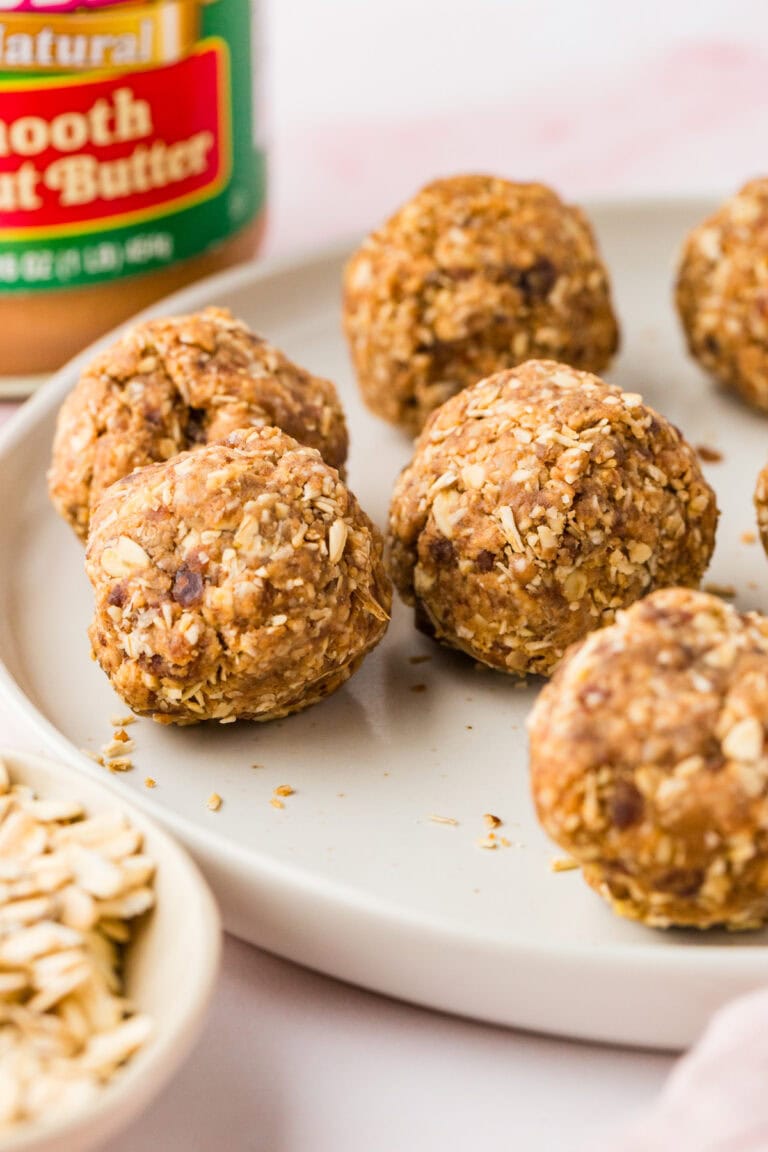 Close-up of several round peanut butter and date protein balls on a white plate, with a jar of smooth peanut butter and a small bowl of oats in the background.
