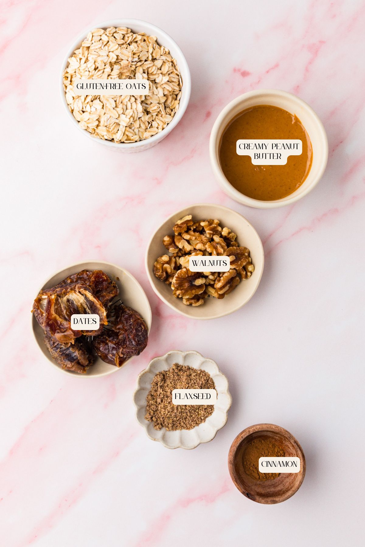 A flat lay of five bowls on a light pink surface contains ingredients for peanut butter and date protein balls: gluten-free oats, creamy peanut butter, walnuts, dates, flaxseed, and cinnamon, each labeled with white tags.