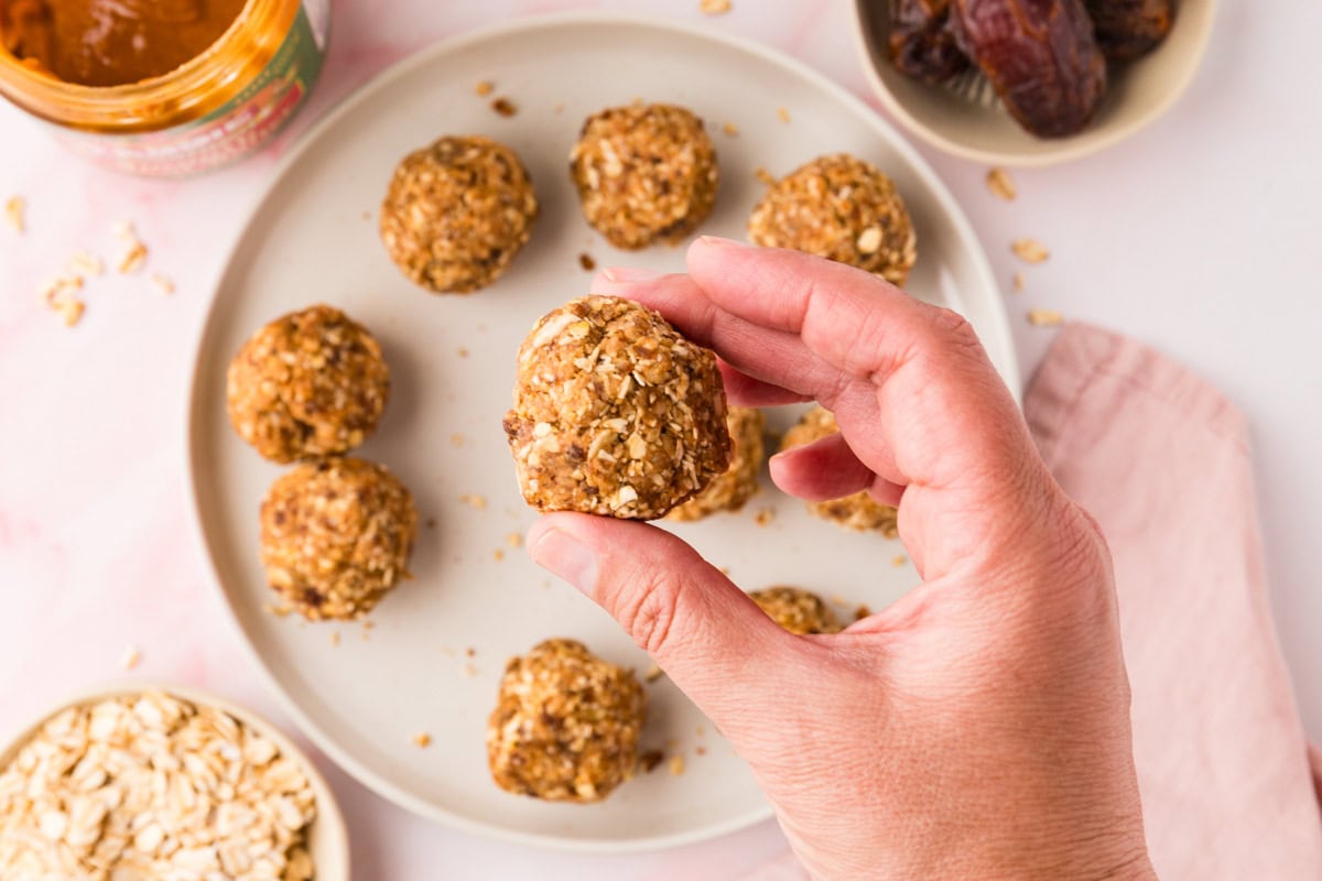 A hand holds an oatmeal energy bite over a plate with more similar bites. Nearby are bowls containing oats and dates, plus a jar of nut butter, all on a light pink surface with a pink napkin.