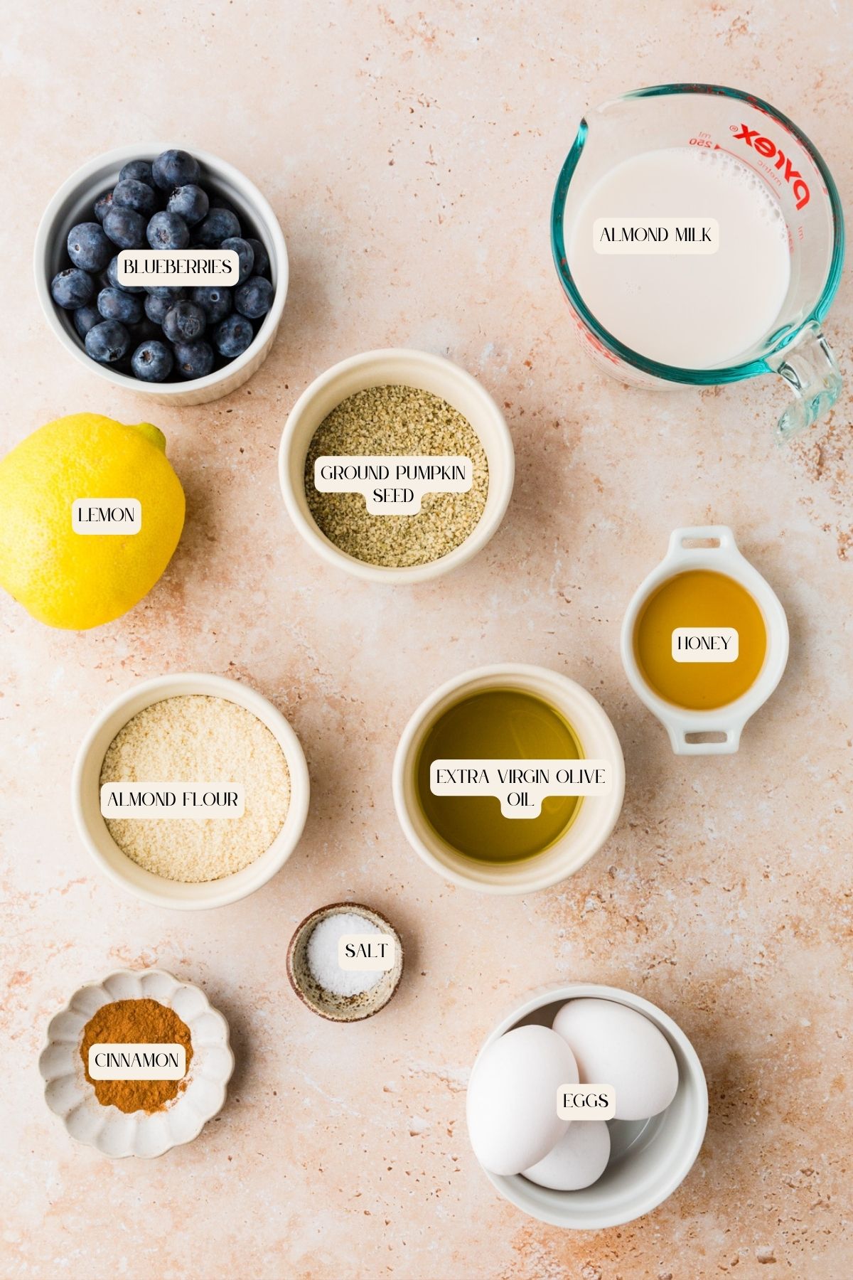 Top-down view of measured baking ingredients for almond flour waffles on a light surface, including blueberries, lemon, ground pumpkin seed, almond flour, almond milk, honey, extra virgin olive oil, salt, cinnamon, and eggs—all labeled.