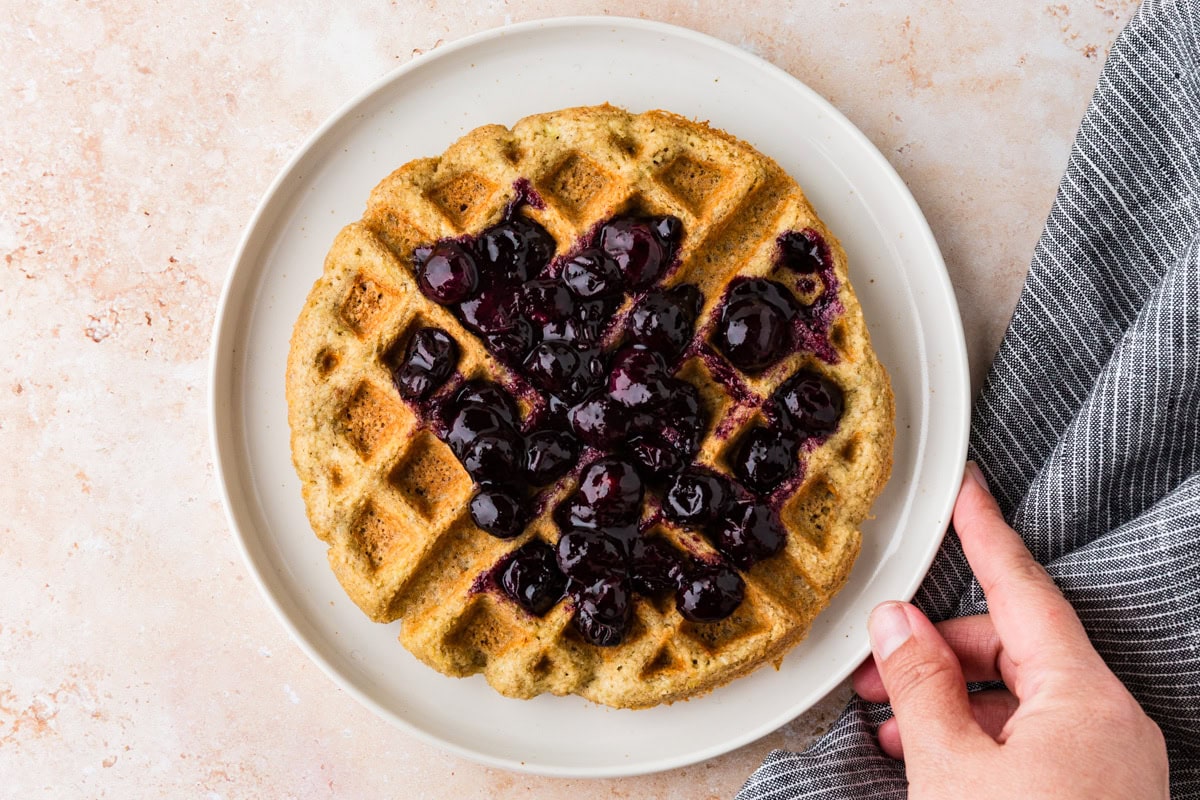 A plate with a single waffle topped with blueberry compote, next to a gray striped cloth. A hand is holding the edge of the plate. The background is a light, speckled surface.