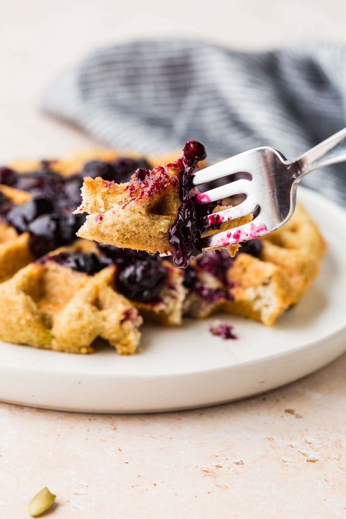 A close-up of a fork holding a bite of almond flour waffle topped with blueberry sauce, with more waffles and sauce on a white plate in the background. A striped towel is partially visible behind the plate.