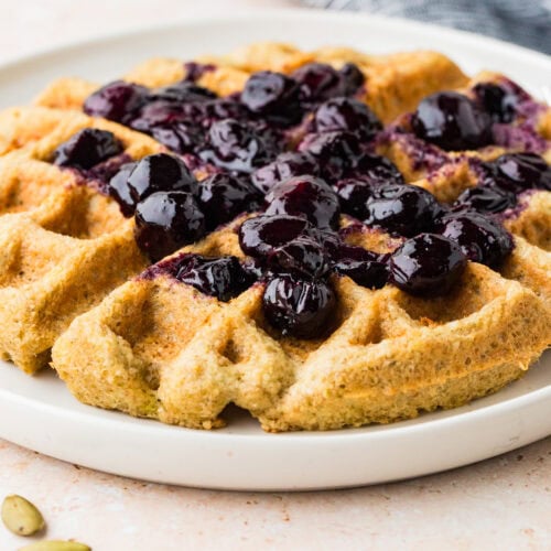A golden-brown almond flour waffle topped with glossy blueberry compote sits on a white plate, with a few cardamom pods nearby. A gray and white checkered cloth is visible in the background.