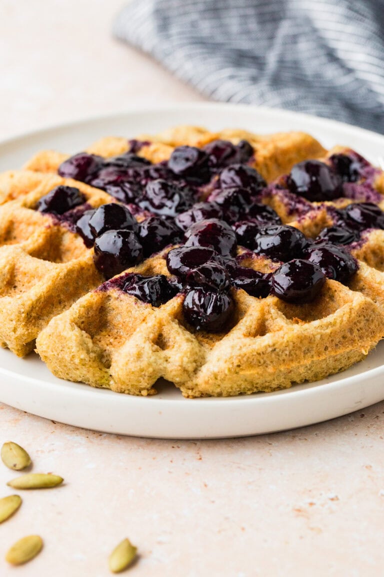 A golden-brown almond flour waffle topped with glossy blueberry compote sits on a white plate, with a few cardamom pods nearby. A gray and white checkered cloth is visible in the background.