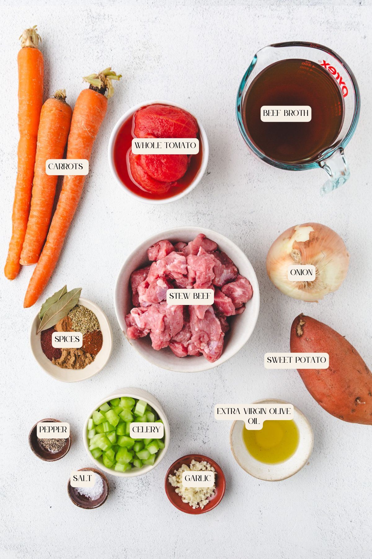Top-down view of labeled Whole30 Beef Stew ingredients: carrots, whole tomato, beef broth, stew beef, onion, sweet potato, extra virgin olive oil, garlic, celery, salt, pepper, and spices on a white surface.