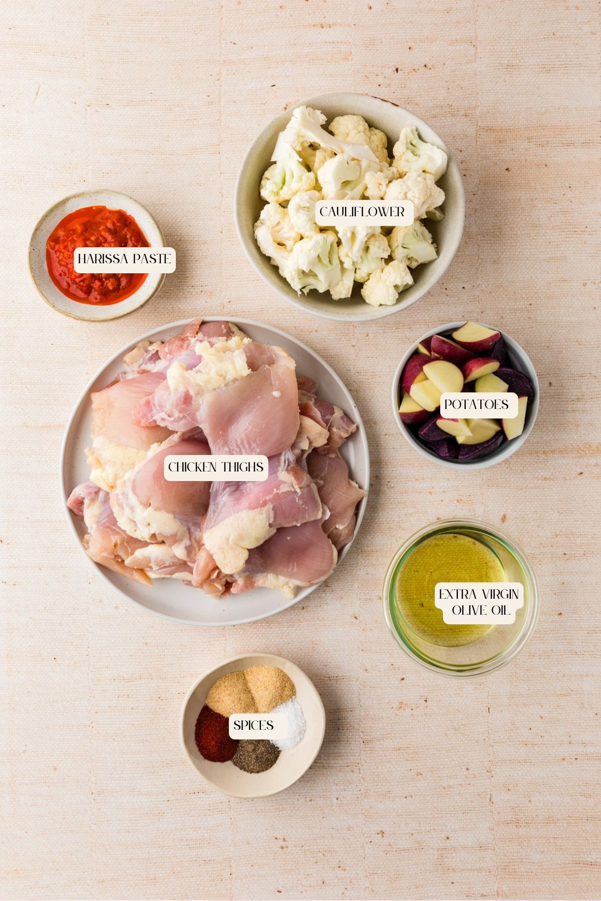 Top-down view of bowls containing harissa paste, cauliflower florets, chopped potatoes, raw chicken thighs for a flavorful harissa chicken recipe, extra virgin olive oil, and assorted spices on a light-colored surface, each labeled with text.