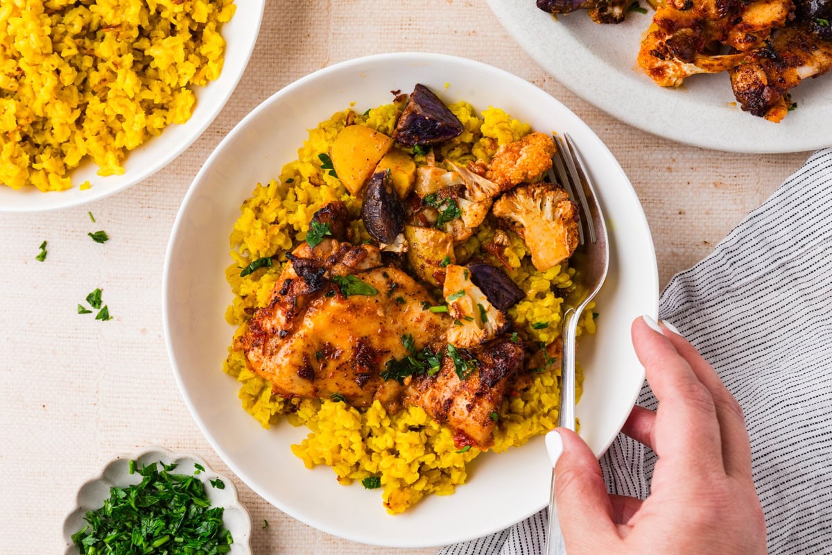 A hand holds a fork in a bowl of yellow rice topped with harissa chicken, roasted vegetables, and fresh herbs. Additional plates of rice, chicken, and herbs are on a light-colored table—perfect for trying a new harissa chicken recipe.