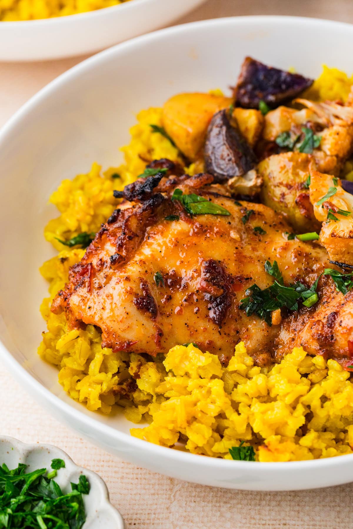 A close-up of a white bowl filled with yellow rice, topped with seasoned, harissa chicken recipe-inspired roasted chicken and garnished with chopped herbs. Pieces of roasted vegetables are also visible.