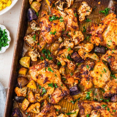 A sheet pan filled with harissa chicken thighs, roasted cauliflower, and chopped potatoes, all seasoned and garnished with fresh herbs. A bowl of yellow rice and a small dish of chopped herbs are nearby—an easy harissa chicken recipe meal.