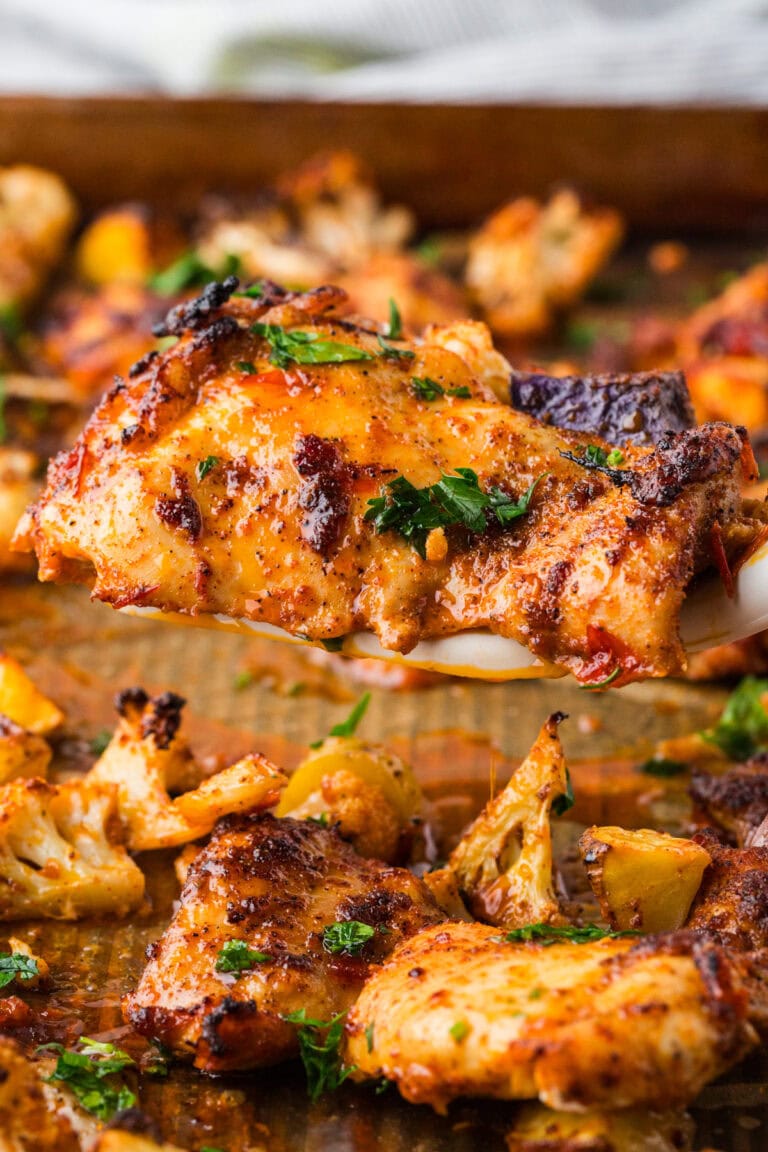 A close-up of a baked, golden-brown harissa chicken recipe garnished with chopped parsley, lifted from a baking sheet with roasted vegetables in the background.