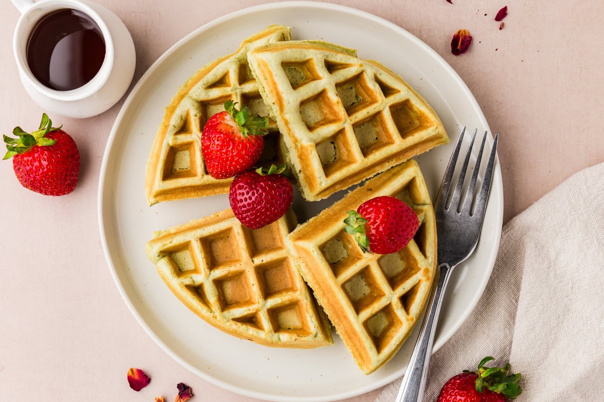 A white plate with four waffle quarters topped with three fresh strawberries, placed next to a fork and knife. A small pitcher of syrup and extra strawberries are nearby on a light tablecloth.
