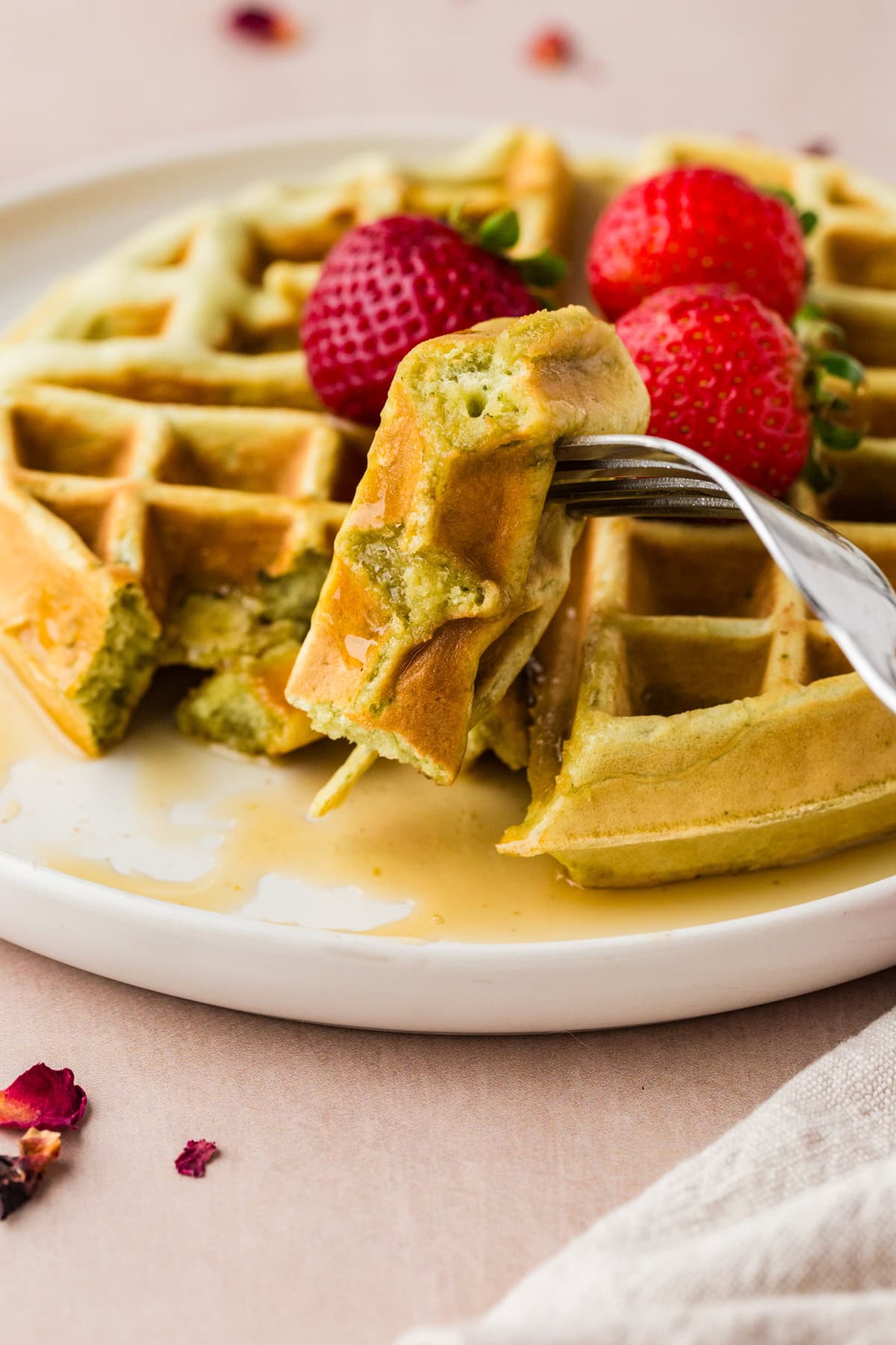 A fork holding a bite of waffle with green filling, with waffles topped with syrup and fresh strawberries on a white plate in the background.