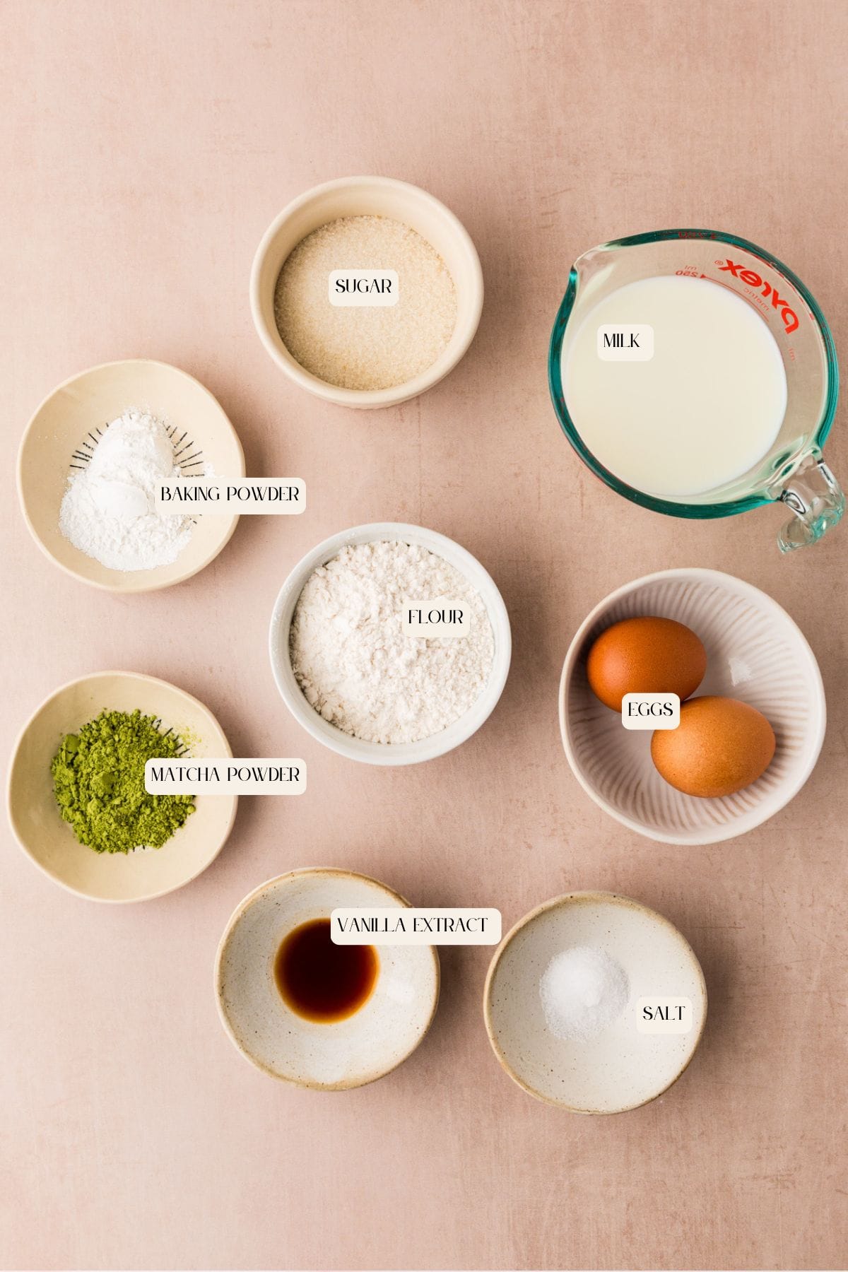 Top-down view of bowls containing labeled ingredients for baking: sugar, milk in a measuring cup, baking powder, flour, eggs, matcha powder, vanilla extract, and salt, arranged on a light pink surface.